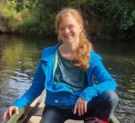 Eine lächelnde Frau sitzt in einem Boot auf einem ruhigen Gewässer. Sie trägt einen blauen Hoodie und genießt die Natur um sie herum.