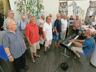 Eine Gruppe älterer Männer steht vor einem Pianisten und singt gemeinsam. Im Hintergrund sind lebendige Tierbilder an der Wand zu sehen.