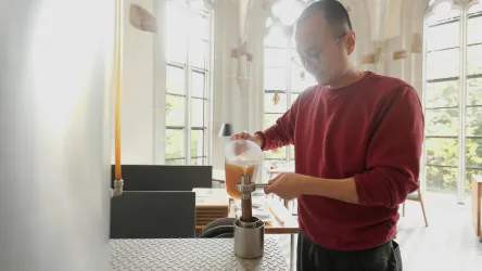 Ein Mann in einem roten Pullover gießt Flüssigkeit in ein Gefäß. Im Hintergrund ist ein heller, moderner Raum mit großen Fenstern zu sehen.