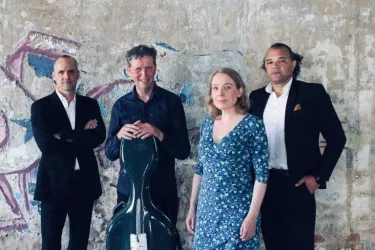 Eine Gruppe von vier Musikern steht vor einer bemalten Wand. Sie haben unterschiedliche Instrumente und tragen elegante Kleidung.