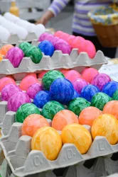 Bunte Ostereier in verschiedenen Farben liegen in einem Karton. Im Hintergrund sind weitere Eier und eine Person mit gestreifter Kleidung zu sehen.