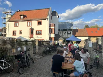 Eine gesellige Veranstaltung im Freien mit Tischen und vielen Menschen. Im Hintergrund sieht man ein charakteristisches Gebäude und Fahrräder.