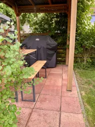 Ein gemütlicher Außenbereich mit einer überdachten Terrasse und zwei Bänken. Im Hintergrund steht ein gecharbter Grill, umgeben von Pflanzen.