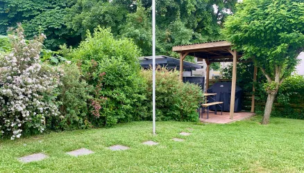 Ein gemütlicher Garten mit viel Grün und bunten Blumen. Im Hintergrund steht eine überdachte Sitzgelegenheit aus Holz.
