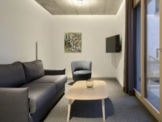 Ein modernes Wohnzimmer mit einem grauen Sofa und einem blauen Sessel. An der Wand hängt ein Kunstwerk, und ein flacher Fernseher ist zu sehen.