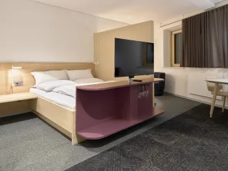 Ein modernes Hotelzimmer mit einem großen Bett, einem Flachbildfernseher und einem kleinen Schreibtisch. Die Einrichtung ist schlicht und stilvoll, mit einem gemütlichen Teppich.