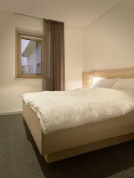 Ein modernes Schlafzimmer mit einem Holzbett und einer weiß-gesteppten Bettdecke. Das Fenster lässt sanftes Licht herein und es gibt einen Vorhang in neutralen Farben.