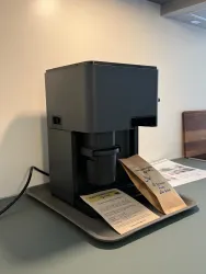 Eine moderne Kaffeemaschine steht auf einem Tisch. Neben der Maschine liegen Kaffeepackungen und eine Anweisung.