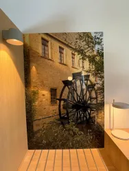 Ein historisches Gebäude mit einem großen Wasserrad im Vordergrund. Die Umgebung ist grün und einladend, was eine ruhige Atmosphäre schafft.
