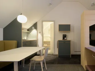 Ein modernes Zimmer mit einer hellen Kommode und einem großen Tisch. Im Hintergrund befinden sich eine Küche und ein Badezimmer.
