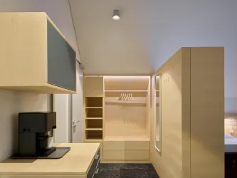 Ein modernes Zimmer mit hellem Holzdekor und minimalistischer Einrichtung. Auf der linken Seite befindet sich eine Kaffeemaschine und ein offener Kleiderschrank.