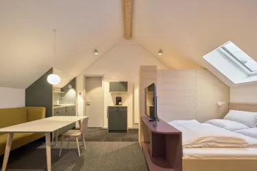 Ein modernes Zimmer mit einem Doppelbett und einem Tisch. Es gibt einen gemütlichen Bereich mit einer kleinen Küchenzeile und viel Tageslicht durch das Dachfenster.