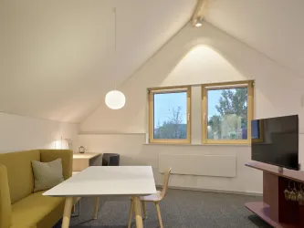 Ein modernes Zimmer mit schrägen Wänden und großen Fenstern. Es gibt einen Tisch, einen Fernseher und eine gemütliche Sitzecke.