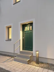Eine moderne Eingangstür in dunklem Grün mit fünf Stufen und einer Außenbeleuchtung. Die Fassade ist hell und schlicht, umgeben von grauen Kieselsteinen.
