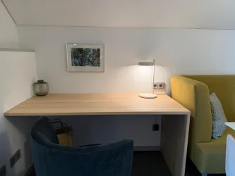 Ein modernes Arbeitszimmer mit einem Holztisch und einem gemütlichen Stuhl. Eine Lampe und ein Bild an der Wand sorgen für eine angenehme Atmosphäre.