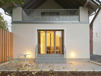 Ein modernes Haus mit einem klaren Eingang und großen Fenstern. Draußen stehen ein kleiner Tisch und Stühle auf einem betonierten Bereich.