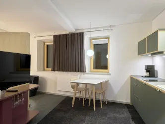 Ein modernes Wohnzimmer mit einem Essbereich und einer Küche. Die großen Fenster lassen viel Licht herein und die Einrichtung ist minimalistisch und stilvoll.