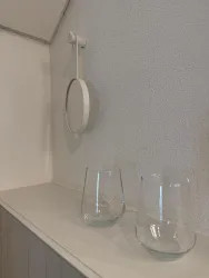 Ein einfacher Tisch mit zwei Gläsern steht an der Wand. An der Wand hängt ein runder Spiegel in einem weißen Rahmen.