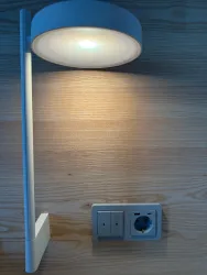Eine moderne Tischlampe leuchtet an einer Holzwand. Darunter befinden sich Steckdosen und Schalter.