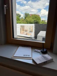 Ein Blick aus dem Fenster zeigt eine grüne Landschaft mit Bäumen und Wolken am Himmel. Auf der Fensterbank liegen Schreibmaterialien und Broschüren.