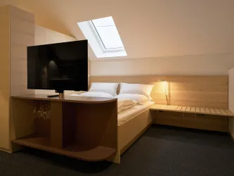 Ein modernes Zimmer mit einem Doppelbett und einem großen Fernseher. Es hat eine gemütliche Atmosphäre mit Holzdesign und Tageslicht von einem Dachfenster.