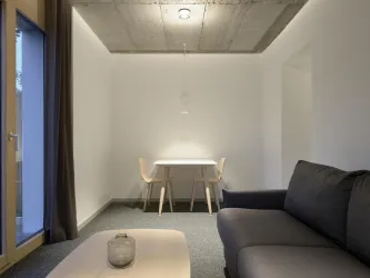 Ein modernes Wohnzimmer mit einem grauen Sofa und einem Tisch mit zwei Stühlen. Die Wände sind hell und das Licht ist sanft.