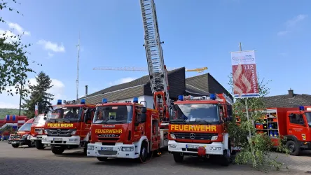 Eine Feuerwehrstation mit mehreren roten Feuerwehrfahrzeugen steht im Vordergrund. Im Hintergrund sind ein hoher Leiternwagen und ein Banner zu sehen.