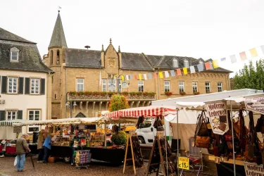Ein Markt mit verschiedenen Ständen und einem historischen Gebäude im Hintergrund. Es sind bunte Wimpel über dem Platz aufgehängt.