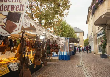 Ein Markt mit verschiedenen Ständen und Waren in einer malerischen Straße. Im Hintergrund sind einige Menschen unterwegs und Bäume sehen aus.