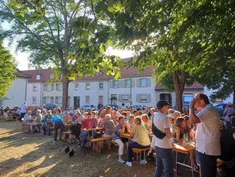 Eine große Menschenmenge sitzt an Tischen unter Bäumen. Es scheint eine fröhliche Veranstaltung im Freien zu sein.