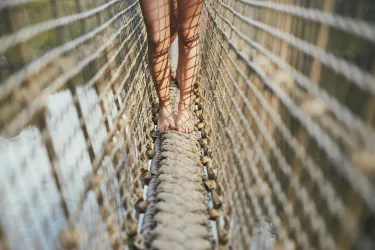 Eine Person steht barfuß auf einer schmalen Hängebrücke. Die Füße sind von einem geschlossenen Netz umgeben, das die Brücke stützt.