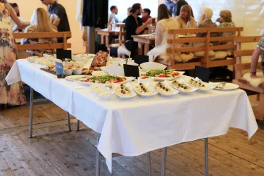 Ein gedeckter Tisch mit verschiedenen Gerichten, darunter Salate und Snacks. Im Hintergrund sind Gäste in einem festlichen Ambiente zu sehen.