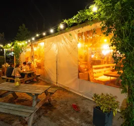 Ein gemütlicher Außenbereich mit einem Zelt, das bei Nacht beleuchtet ist. Menschen sitzen an einem Tisch und genießen die angenehme Atmosphäre.