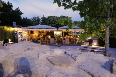 Ein gemütliches Restaurant im Freien mit überdachten Sitzbereichen und warmem Licht. Umgeben von Bäumen und einer einladenden Atmosphäre.