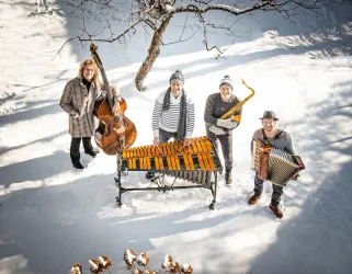 Eine Gruppe von vier Musikern steht im Schnee. Sie spielen verschiedene Instrumente und tragen warme Kleidung.