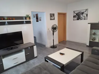 Ein modernes Wohnzimmer mit einem Fernseher, einem Couchtisch und einer bequemen Couch. An der Wand hängen Bilder und es steht ein Ventilator im Raum.