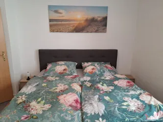 Ein gemütliches Schlafzimmer mit einem Doppelbett und bunten Blumenbettwäsche. An der Wand hängt ein Bild von einem Strand bei Sonnenuntergang.