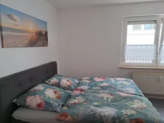 Ein gemütliches Schlafzimmer mit einem großen Bett und bunten Bettbezügen. An der Wand hängt ein Bild von einem Strand bei Sonnenuntergang.