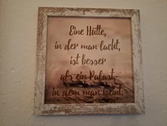 Ein gerahmter Spruch auf einem sanften Hintergrund. Der Text besagt, dass eine Hütte, in der gelacht wird, besser ist als ein Palast.