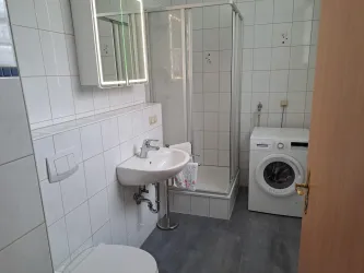 Ein modernes Badezimmer mit Dusche, Waschbecken und Waschmaschine. Die Wände sind weiß gefliest und der Boden ist grau.