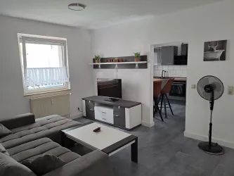 Ein modernes Wohnzimmer mit einem grauen Sofa und einem Fernseher. Der Raum ist hell und freundlich, mit Zugang zur offenen Küche.
