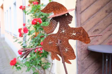 Eine rostige Skulptur einer Hexe mit einem Hut, die auf einem Besen reitet. Im Hintergrund blühen rote Rosen und es gibt eine ruhige Gebäudefassade.