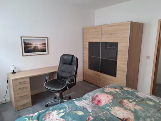 Ein modernes Schlafzimmer mit einem bequemen Schreibtisch und einem schwarzen Bürostuhl. Im Hintergrund steht ein geräumiger Kleiderschrank und ein Bild an der Wand.