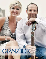 Ein lächelndes Paar sitzt zusammen, der Mann hält eine Gitarre. Im Hintergrund ist ein unscharfer, heller Raum zu sehen.