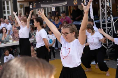 Eine Tanzgruppe führt eine Aufführung vor einem Publikum auf. Die Tänzerinnen tragen weiße T-Shirts und schwarze Hosen und zeigen energievolle Bewegungen.