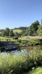 Eine ruhige Landschaft mit einem kleinen Fluss und einer Hängebrücke. Im Hintergrund sind grüne Hügel und Bäume zu sehen.
