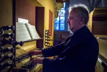 Ein Mann spielt Orgel in einer Kirche. Im Hintergrund sind Notenblätter sichtbar.