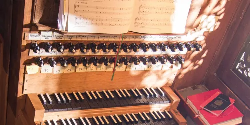 Eine Orgel mit vielen Reglern und Tasten ist im Bild zu sehen. Darüber liegt ein Notenblatt mit Musiknotation.