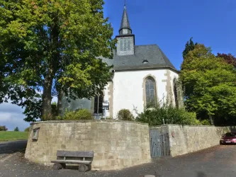 Eine kleine Kirche mit einem spitzen Turm und einer umgebenen Mauer. Im Vordergrund steht eine Bank und es sind einige Bäume zu sehen.
