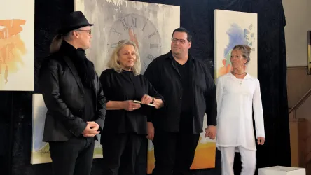 Vier Personen stehen auf einer Bühne, umgeben von Kunstwerken. Sie scheinen an einer Präsentation oder Veranstaltung teilzunehmen.
